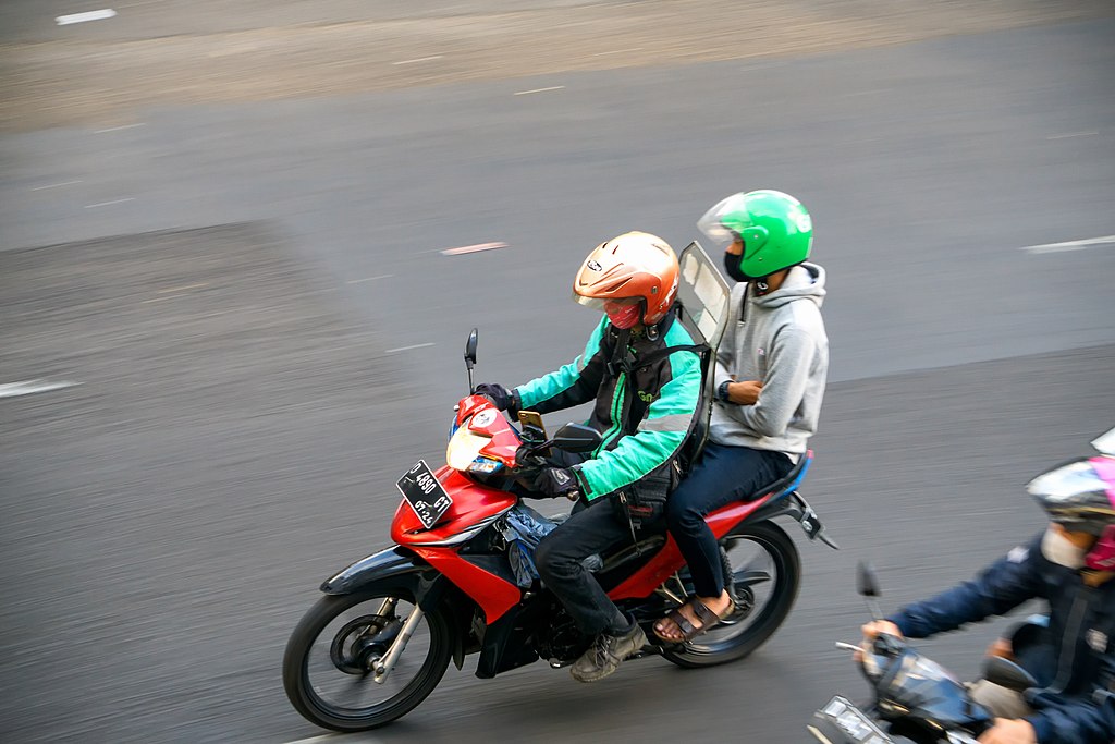 Dua orang di atas motor merah di jalan raya yang ramai. Orang di depan memakai helm putih dan jaket hitam, dan memegang helm merah putih di pangkuannya. Orang di belakang memakai helm hijau dan hoodie abu-abu. Motor merah memiliki aksen hitam dan kotak hitam di belakangnya.